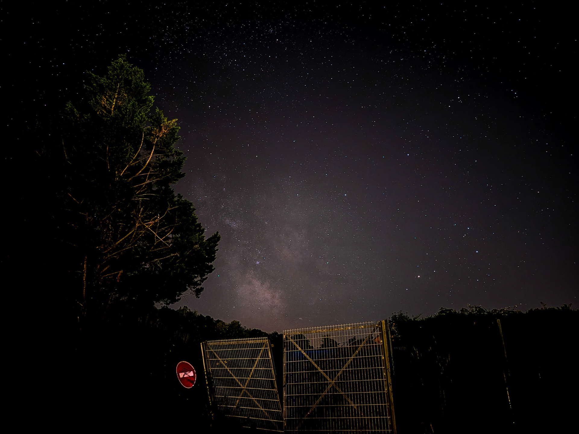 Milky Way arching above a gate and lone tree at night